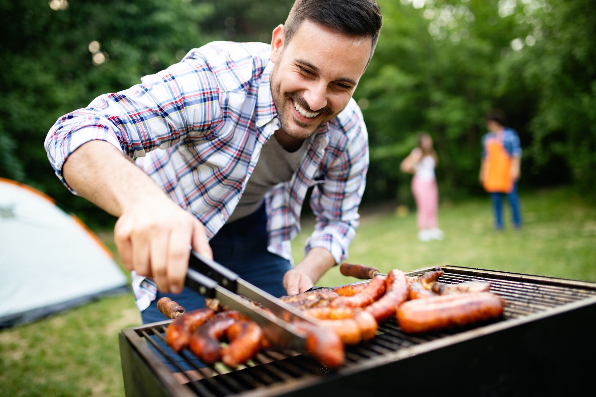 Ein Mann der draußen im garten Bratwürste grillt und im Hintergrund reden zwei Personen